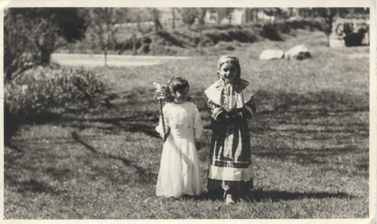 Plano entero de dos niñas disfrazadas. A la izquierda una está disfrazada de hada y a la derecha está disfrazada de gitana. Se encuentran paradas sobre el pasto, en un sector abierto no identificado.
