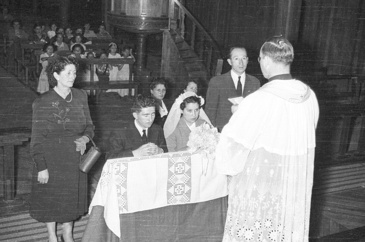 Plano entero de un sacerdote dirigiéndose a la pareja de novios, los cuales se encuentran arrodillados en el reclinatorio y a sus padrinos que están de pie junto a ellos. En el fondo, hay un grupo de Mujer y Niñas sentadas en las bancas durante la misa del sacramento de matrimonio en el interior de una iglesia.