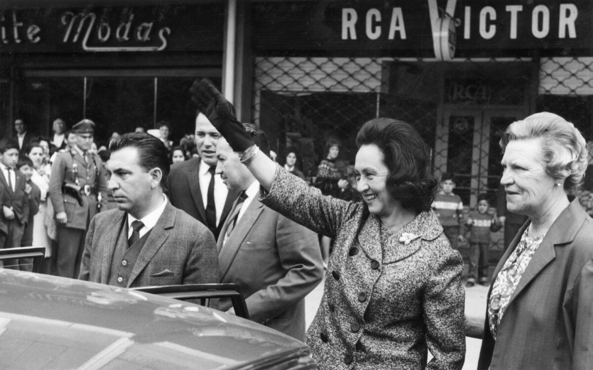 plano general de Eva Sámano, saludando a las personas antes de subir a un automóvil. Del brazo la acompaña Olga Boettcher. En el fondo, hay un grupo numeroso de Niños, Mujeres, Hombres y Niñas parados en el frontis de unas tiendas comerciales, durante la visita de la primera dama a la ciudad de La Unión