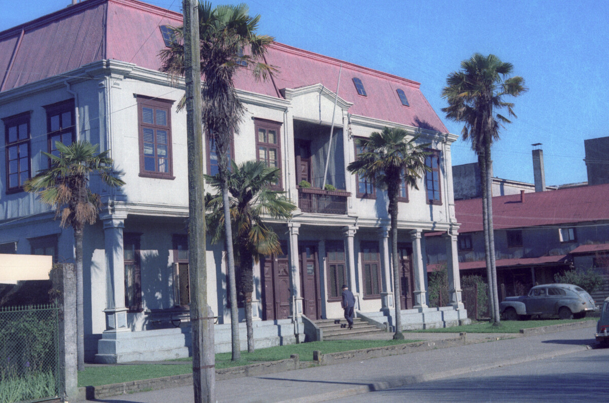 Plano general de una gran casa de tres niveles con un antejardín con palmeras frente a una calle pavimentada