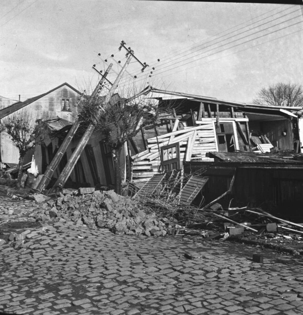 Reproducción de fotografía. plano general de un tendido eléctrico caído sobre los escombros de una casa tras el terremoto del 60' en la ciudad de Valdivia