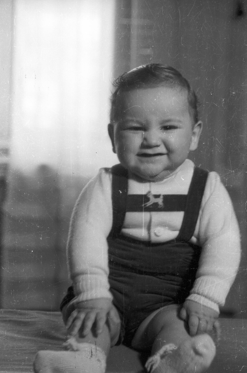 Retrato de plano entero de un Bebé con jardinera, sonriendo mientras posa de frente, sentado en una cama