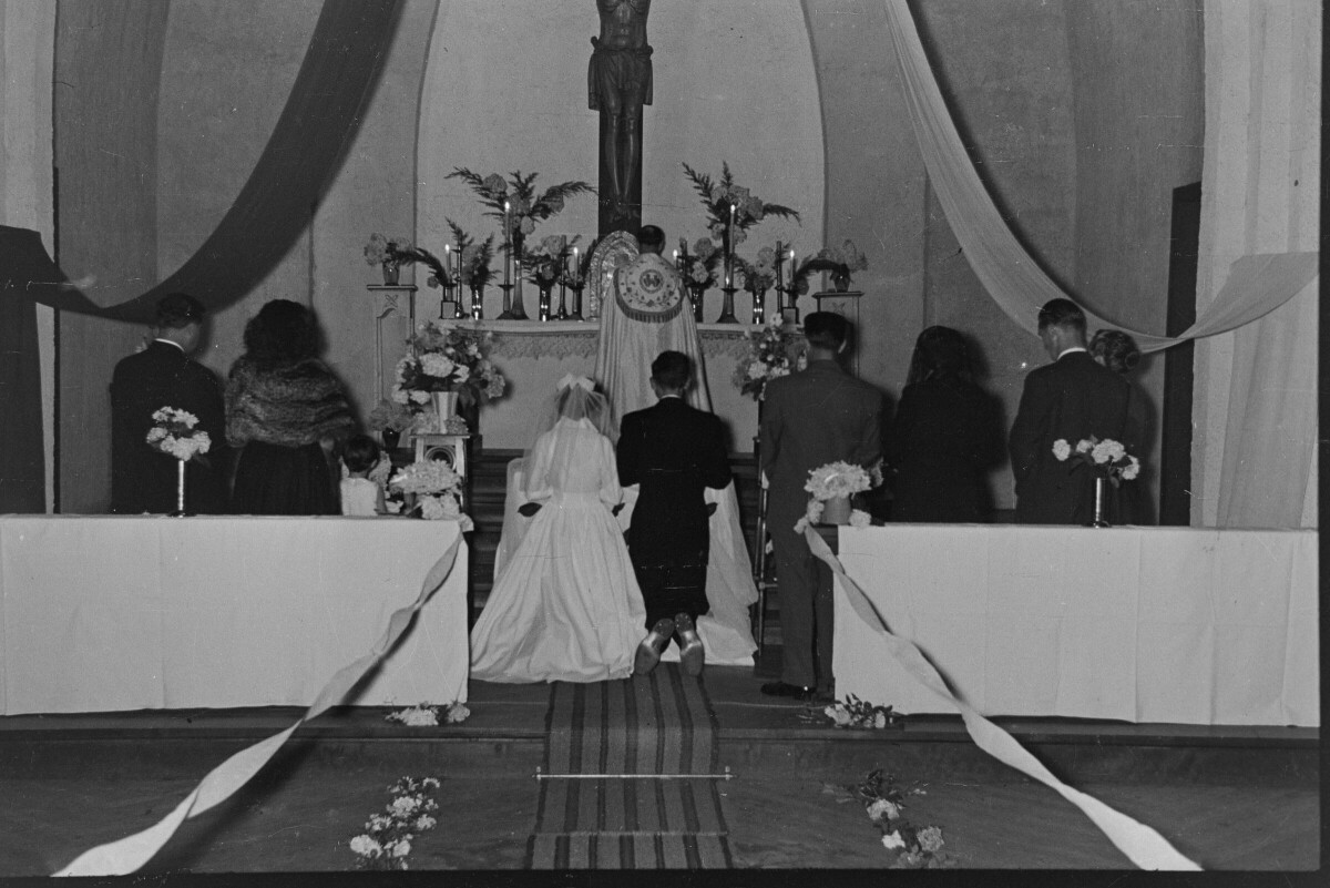 plano general de una pareja de novios, hincados sobre un reclinatorio en el altar. Ellos, al igual que sus padrinos de bodas que los acompañan, se encuentran de espaldas hacia la cámara. En el fondo, un sacerdote se encuentra parado frente a las velas del altar. Se encuentran en el interior de la parroquia San José lugar donde se está llevando a cabo la misa de celebración de su casamiento