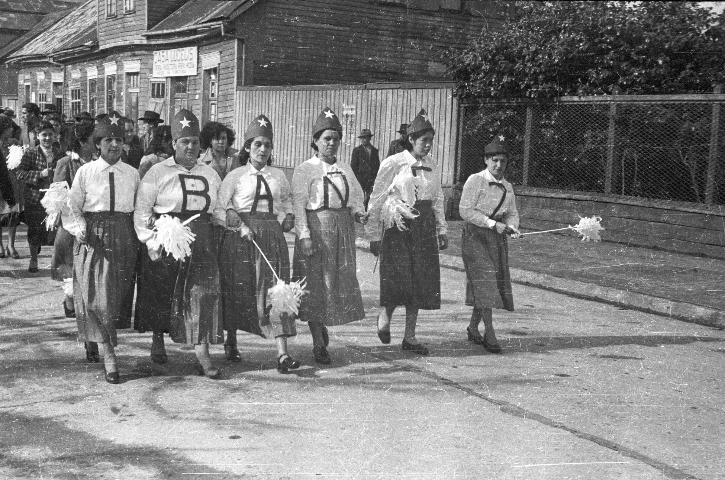 Plano entero de un grupo de mujeres desfilando, cada una lleva una letra en sus poleras las cuales forman la palabra Ibáñez, llevan puesto además un gorro con una estrella y pompones en sus manos, detrás de ellas viene un grupo de hombres y mujeres desfilando por una calle durante la campaña presidencial de Carlos Ibáñez del Campo en La Unión