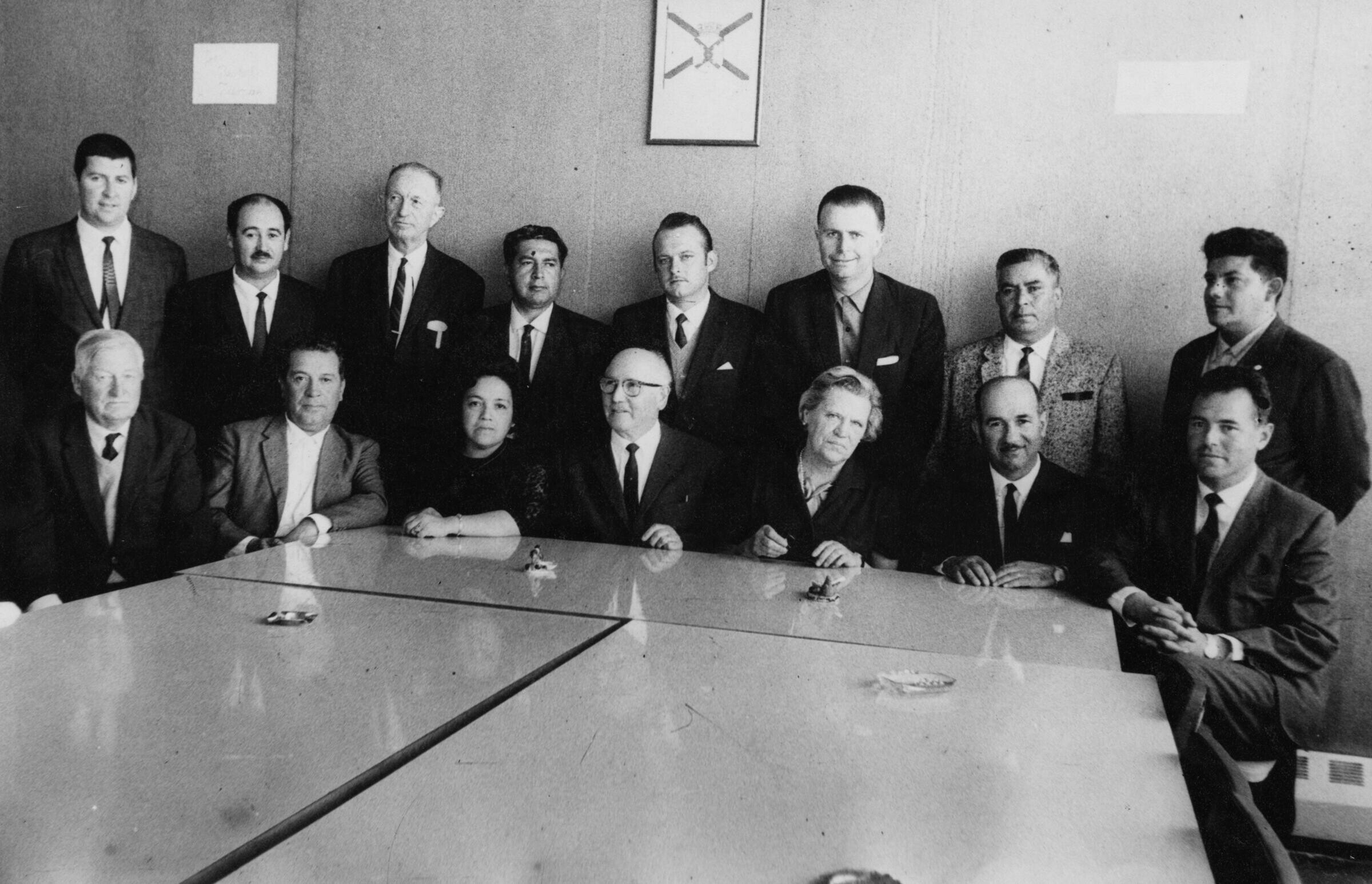 Retrato grupal de plano entero de Hombres y dos mujeres posando reunidos tras una mesa. En primera fila, la segunda mujer sentada es Olga Boettcher. Se encuentran en una infraestructura no identificada
