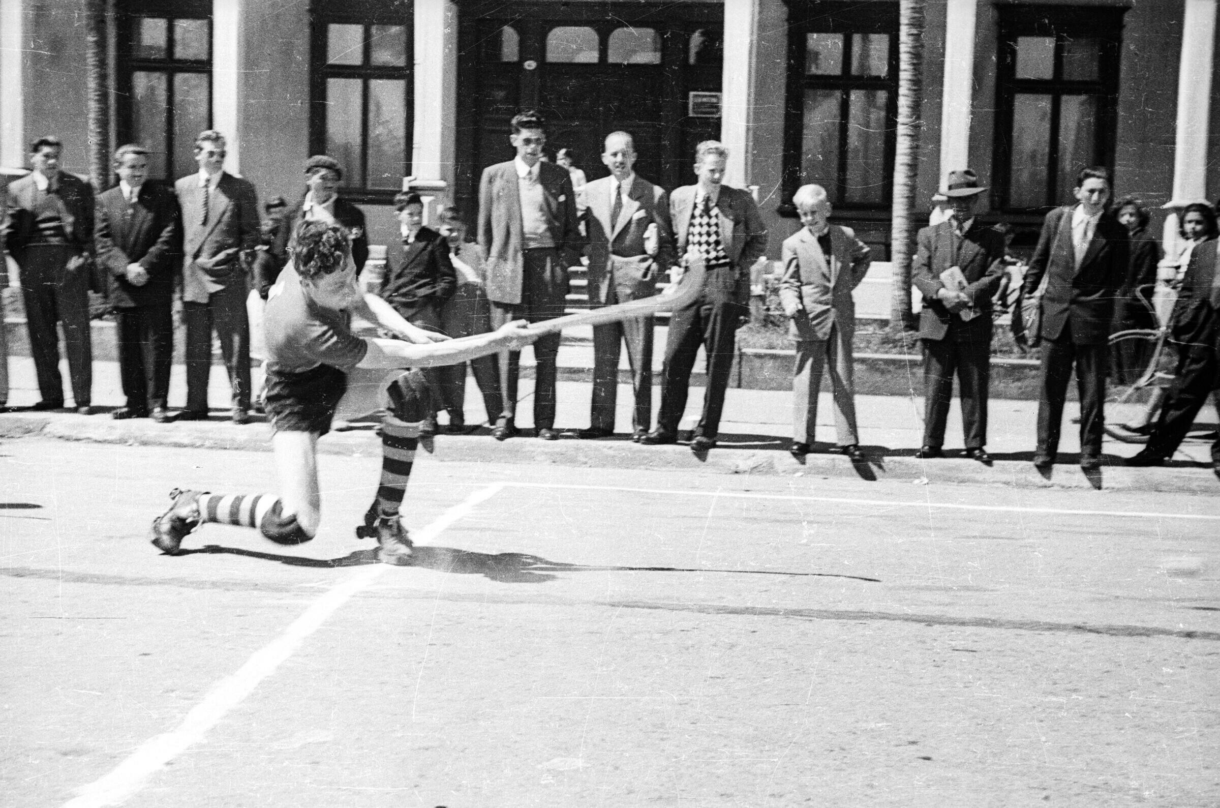 plano general de un hombre tras haber golpeado una pelota con su palo de hockey durante un partido, en el fondo hay Hombres y Niños observándolos mientras juegan en la calle en las afueras de un edificio