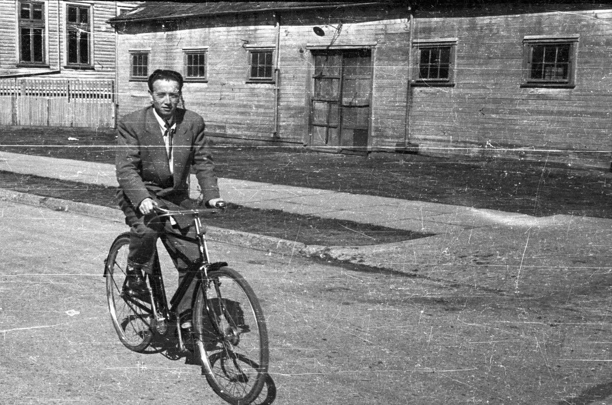 Plano entero de un hombre andando en una bicicleta camello, vestido de traje por una calle pavimentada