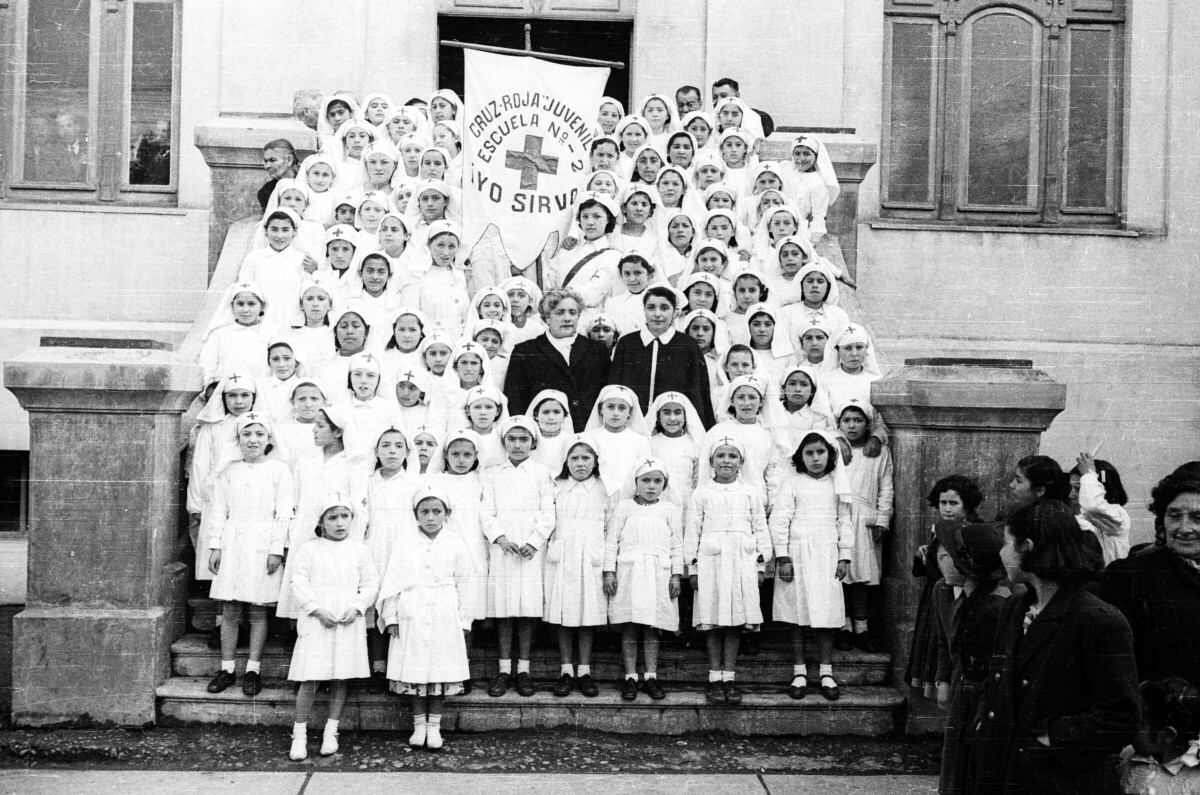 Retrato grupal de plano entero de un grupo de Niñas y dos Mujeres pertenecientes a la Cruz Roja Juvenil de la Escuela Nº2, las niñas están vestidas de blanco y en la cabeza llevan una túnica con una cruz en la frente, entre ellas tienen puesto el pendón de la agrupación. están posando en la escalera de acceso de un colegio.
