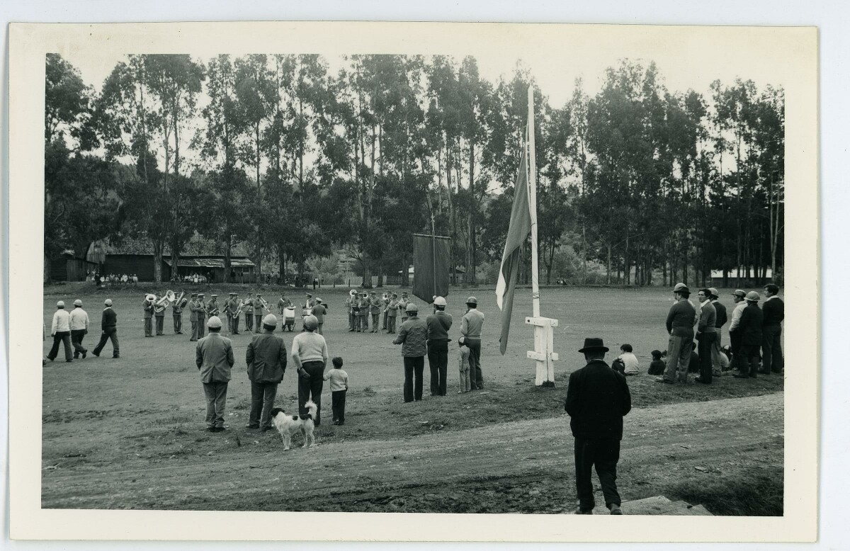 plano general de una orquesta de carabineros, tocando sus instrumentos a la mitad de un recinto, mientras un grupo de mineros e hijos, los observa. Está actividad se lleva a cabo en los recintos de la compañía San Pedro de Catamutún, La Unión