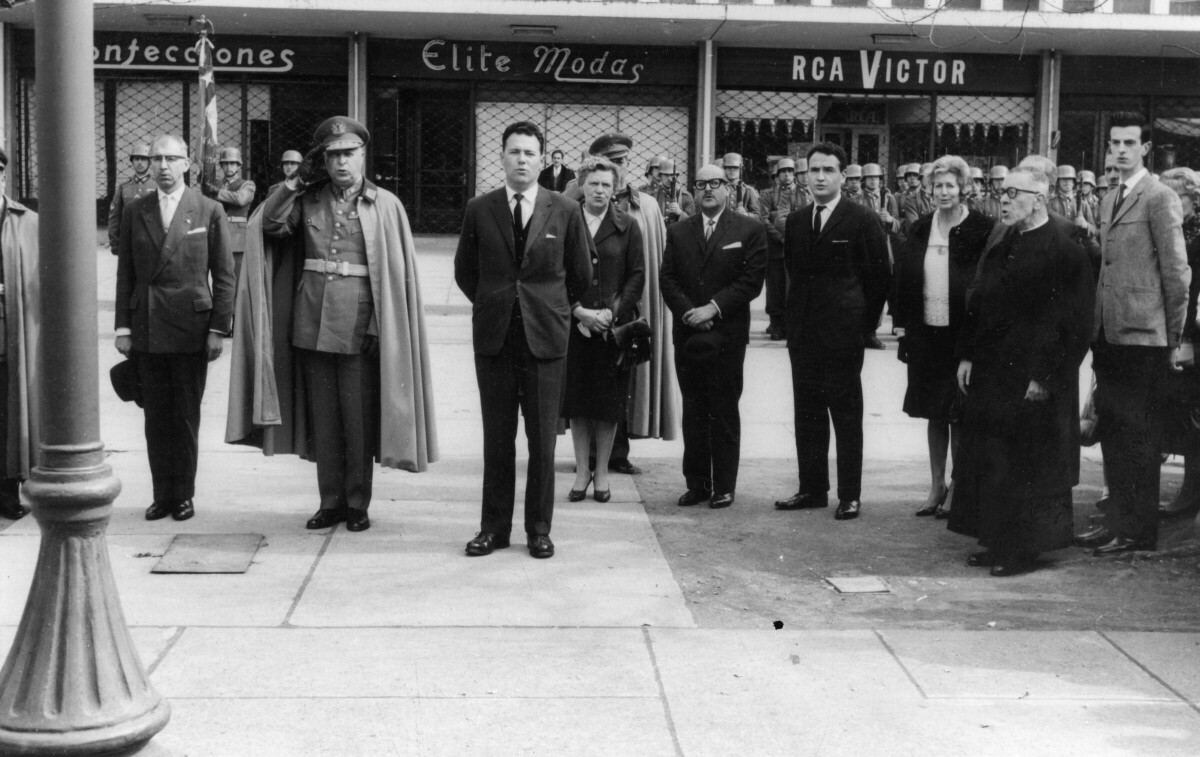 plano general de un grupo de Hombres, Mujeres, un militar y un sacerdotes reunidos sobre la vereda. La primera mujer de izquierda a derecha es Olga Boettcher. En el fondo, se encuentra un grupo de militares formados sobre la calle, durante la visita de la escuela militar Bernardo O'Higgins a la ciudad de La Unión