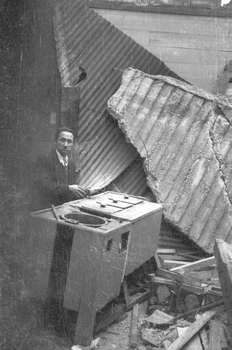 Plano entero de hombre posando junto a maquina de confites entre los escombros de infraestructura dañada por el terremoto del 60'.
