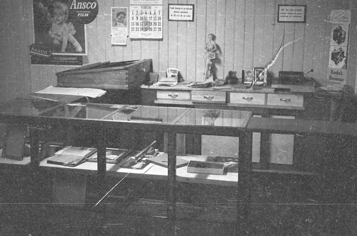 plano general de un mostrador con fotografías y marcos de fotos en su interior. En la pared del fondo hay dos letreros que dicen Ansco, Film' y Kodak Brownie'. También hay un calendario que indica el mes de febrero que dice Sud América'. está vista interior corresponde al interior de un estudio fotográfico