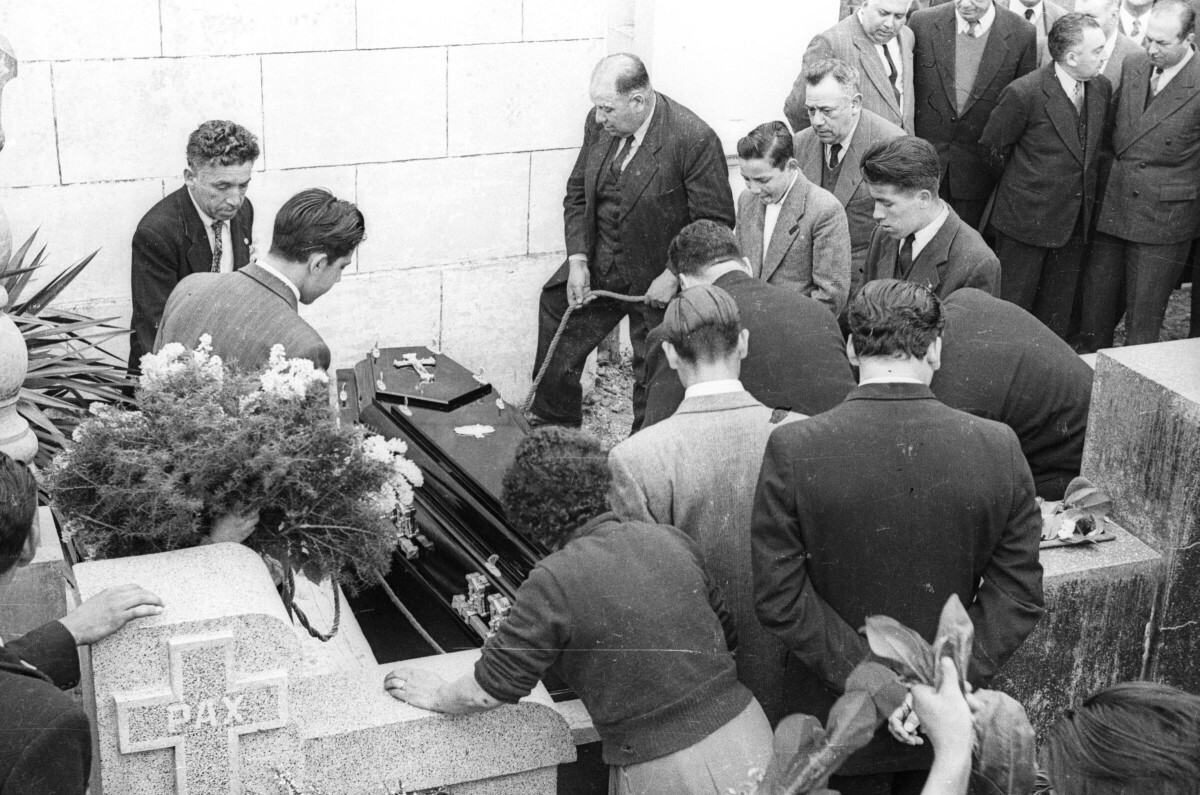 Plano entero de un grupo de hombres vestidos de traje, descendiendo un féretro en su tumba. A su alrededor los acompaña un Niño , una mujer y un grupo de hombres durante el funeral en el interior de un cementerio no identificado