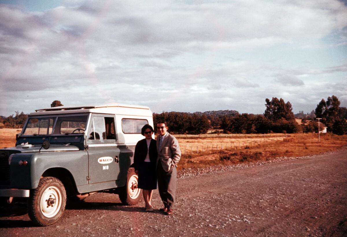 La fotografía muestra a dos personas de pie (Yoyi Koda y una acompañante) junto a un vehículo tipo todoterreno, ubicado en un camino de tierra dentro de un paisaje campestre. A la izquierda de la imagen, el vehículo es de color azul y tiene un techo blanco con el texto visible 'RALCO M8' cerca de la puerta del pasajero. Las personas, un hombre y una mujer, visten ropa de corte formal; él lleva un traje claro y ella una falda y chaqueta oscuras. El fondo de la imagen se compone de un campo con pasto, cercado por árboles y arbustos. El cielo está mayormente nublado, con algunas áreas despejadas que dejan ver un paisaje amplio y abierto.