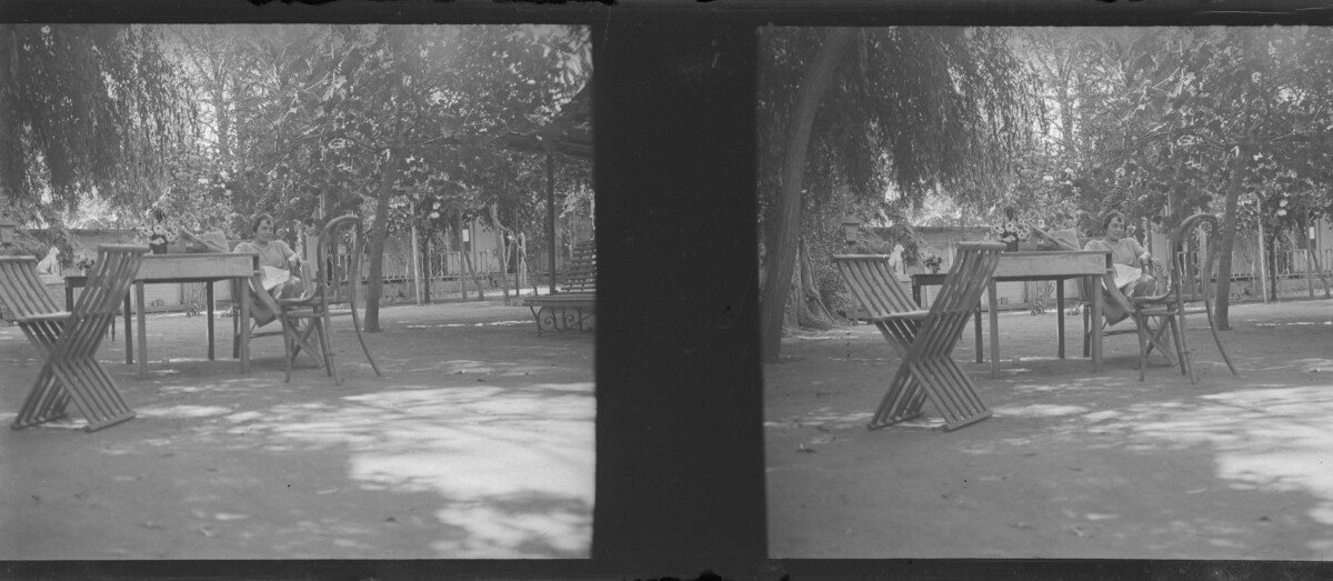 Imagen estereoscópica. Retrato de plano entero de mujer con vestido sentada junto a una mesa, con los pies reposando sobre otra silla, en un lugar rodeado de casas