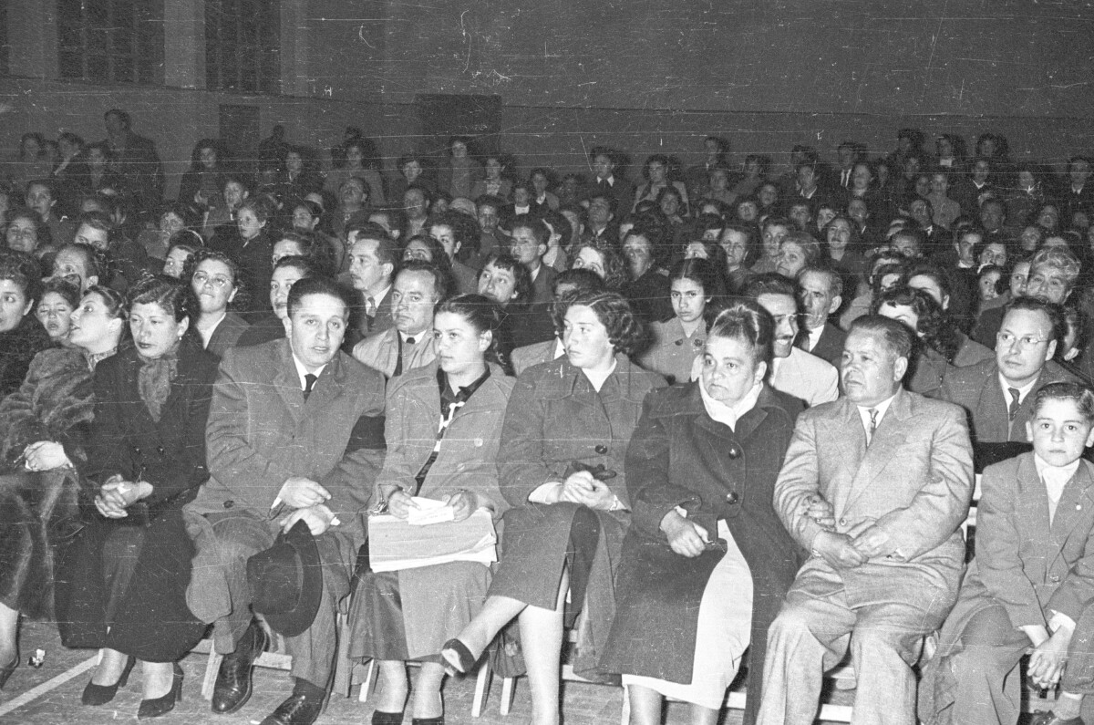 plano general de un grupo de Mujeres, Hombres, Niños y Niñas sentados en el interior del gimnasio observando hacia el escenario las presentaciones de los estudiantes durante una jornada de actividades escolares en el gimnasio de un colegio