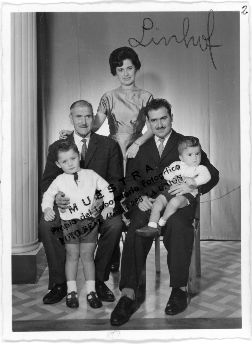 Retrato de estudio de Plano entero de un hombre mayor, posando sentado con un niño parado entre sus piernas. En la silla de al lado, hay un hombre vestido de Traje que está posando con un niño sentado sobre sus piernas y por detrás hay una mujer posando parada.