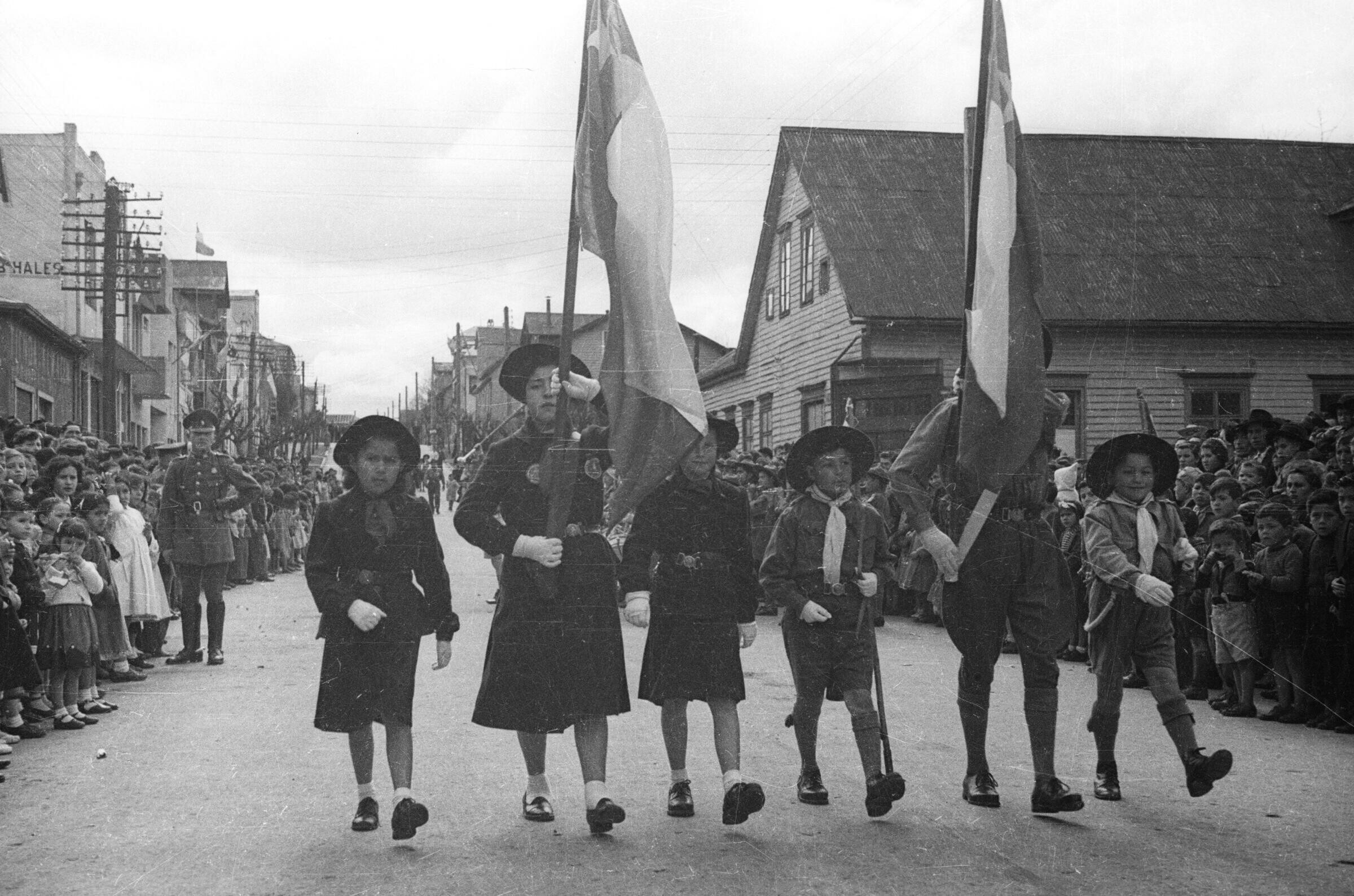 Plano entero de un grupo de Niños pertenecientes a la orquesta sinfónica de la brigada de scout, tocando sus instrumentos formados en hileras, detrás de un Niño , quién los dirige moviendo la guaripola. Se encuentran ubicados en la intersección de dos calles durante un desfile realizado en la ciudad de La Unión