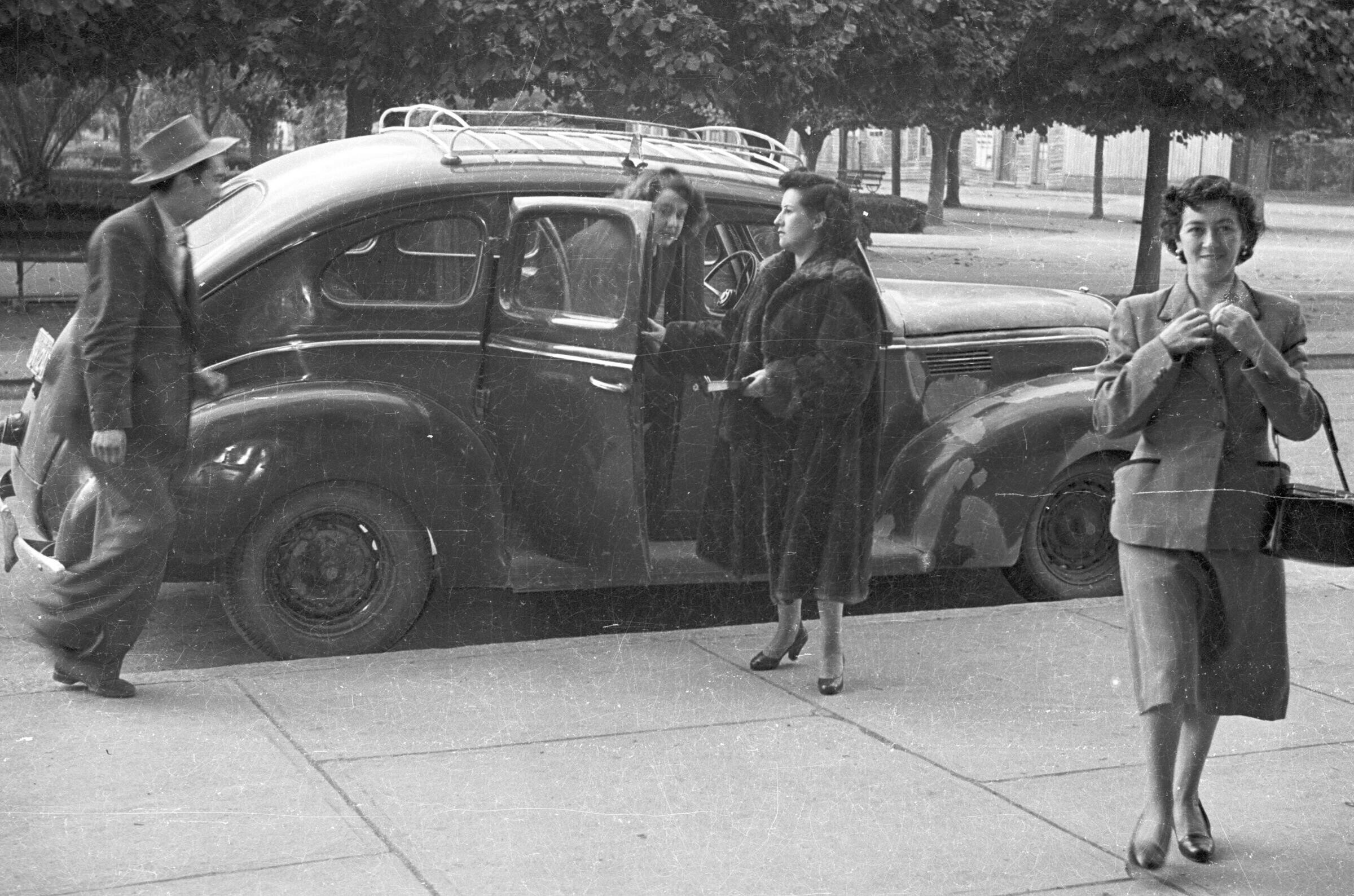 Plano entero de una mujer vestida de formalmente mirando hacia la cámara. detrás de ella, otras dos mujeres que visten con abrigo, descienden de un auto estacionado, mientras un hombre con sombrero se aproxima a ellas. El auto se encuentra estacionado a un costado de la vereda, en el frontis de la parroquia San José
