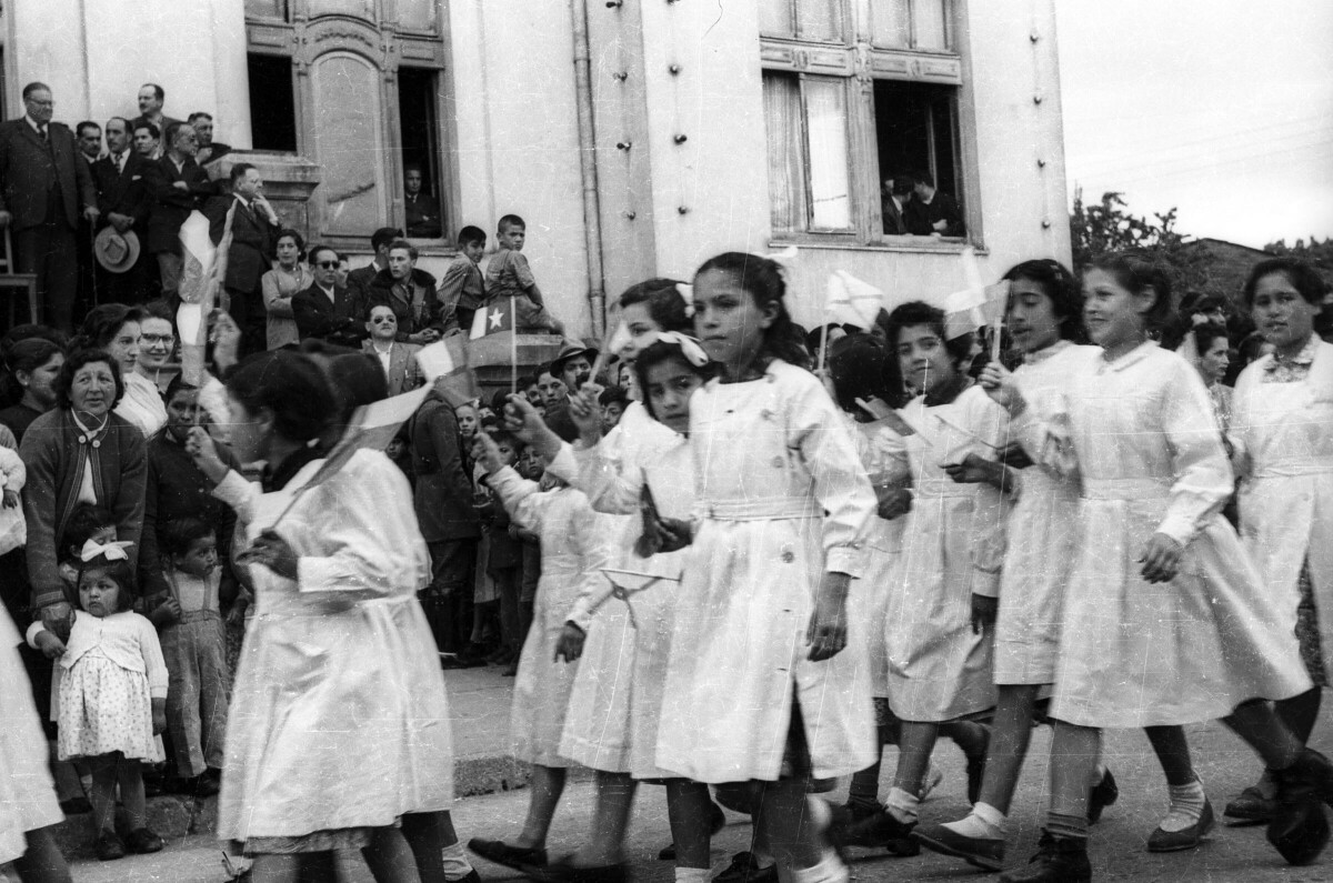 plano general de un grupo de niñas con delantal, desfilando formadas en hileras, con una bandera en las manos. En el fondo, un grupo de personas y Carlos Vogel se encuentran observándolas en el frontis de la municipalidad