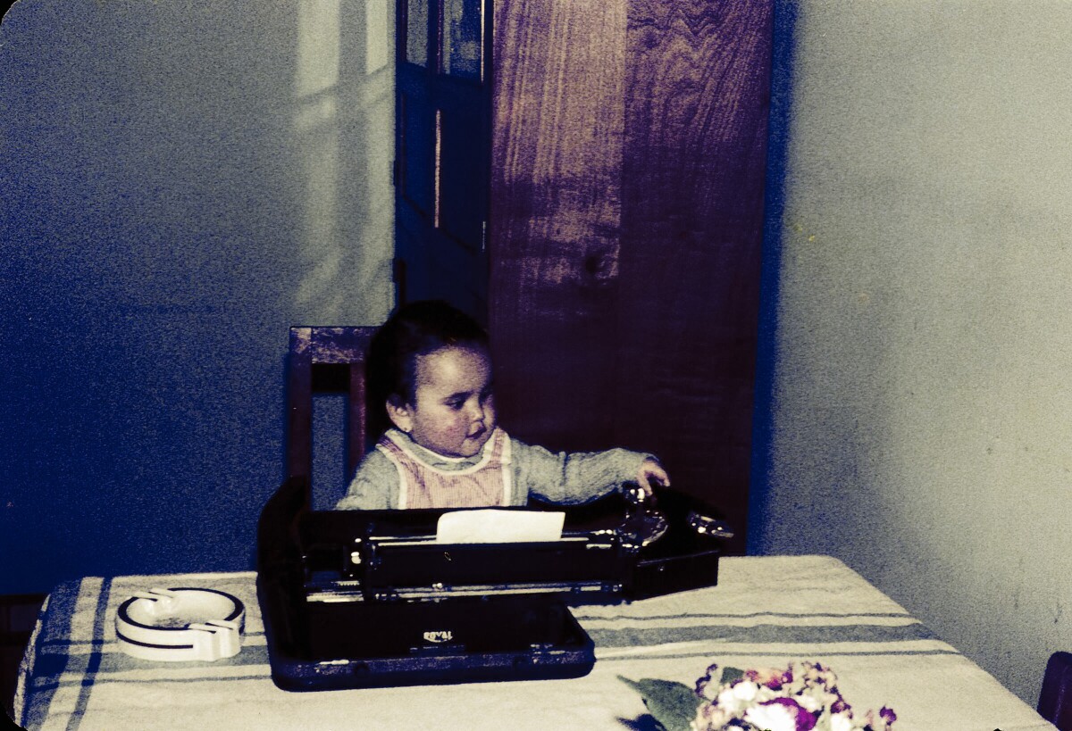 Retrato de plano medio de niña de temprana edad sentada junto a una mesa con una máquina de escribir y un teléfono en lugar no identificado