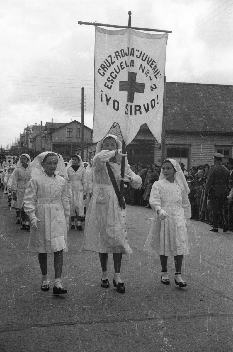 -Plano entero de tres Niñas desfilando formadas en una hilera. La Niña que está ubicada en el medio, va levantando un pendón el cual tiene escrito -- Cruz Roja Juvenil Escuela Nº2 ¡Yo Sirvo!--. A lo largo de toda la calle, le siguen otro grupo de Niñas, también pertenecientes de la cruz roja. Todas utilizan una vestimenta de color blanco, la cual consiste en un Vestido, guantes y un velo. A un costado de la calle hay un grupo de Hombres, Niños, Niñas y Mujer observándolas transitar durante el desfile realizado por una de las calles de la ciudad de La Unión-