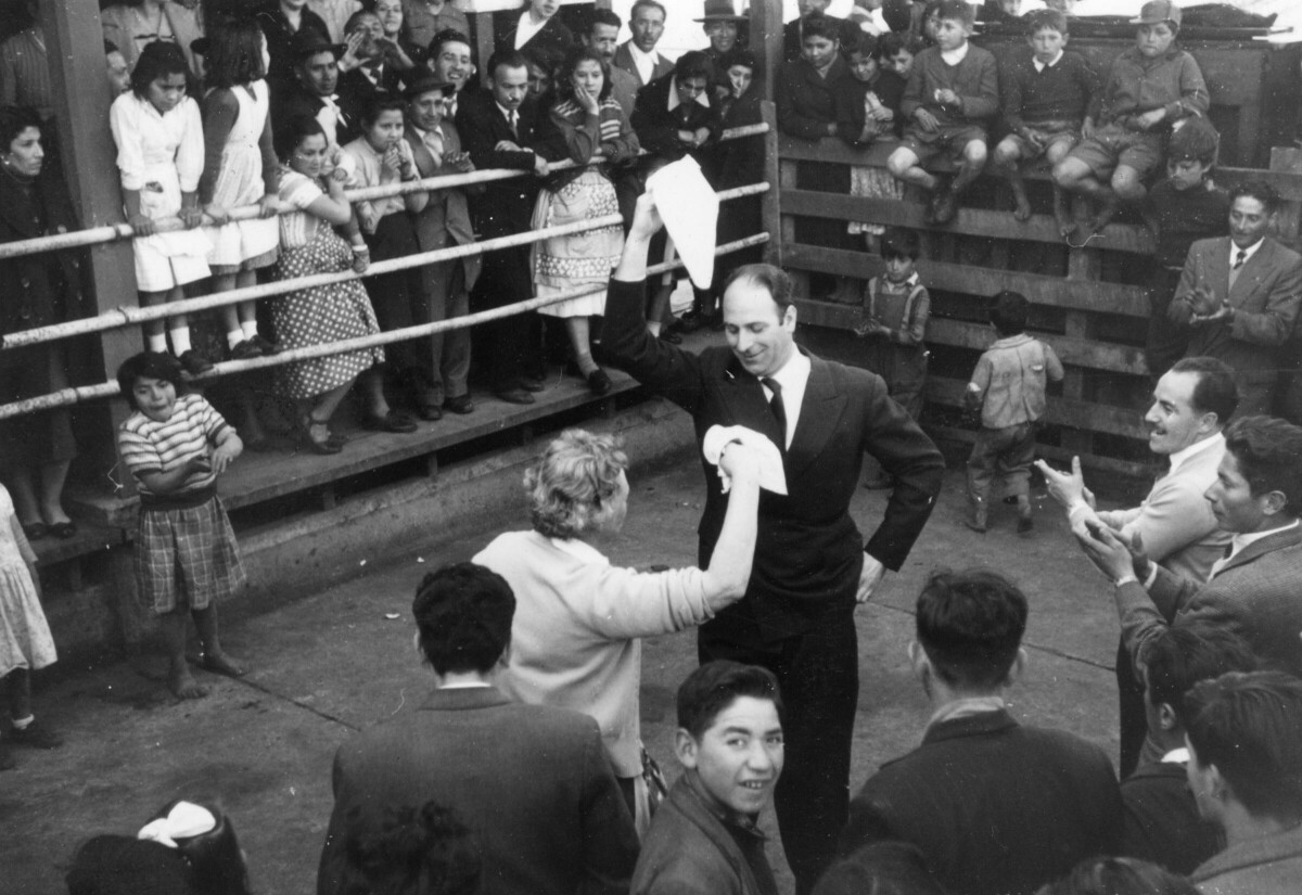plano general de Olga Boettcher, bailando un pie de cueca junto a un hombre vestido de Traje mientras ambos agitan sus pañuelos. A su alrededor, un grupo numeroso de Niñas, Niños, Hombres y Mujeres los acompañan en el interior de un recinto no identificado