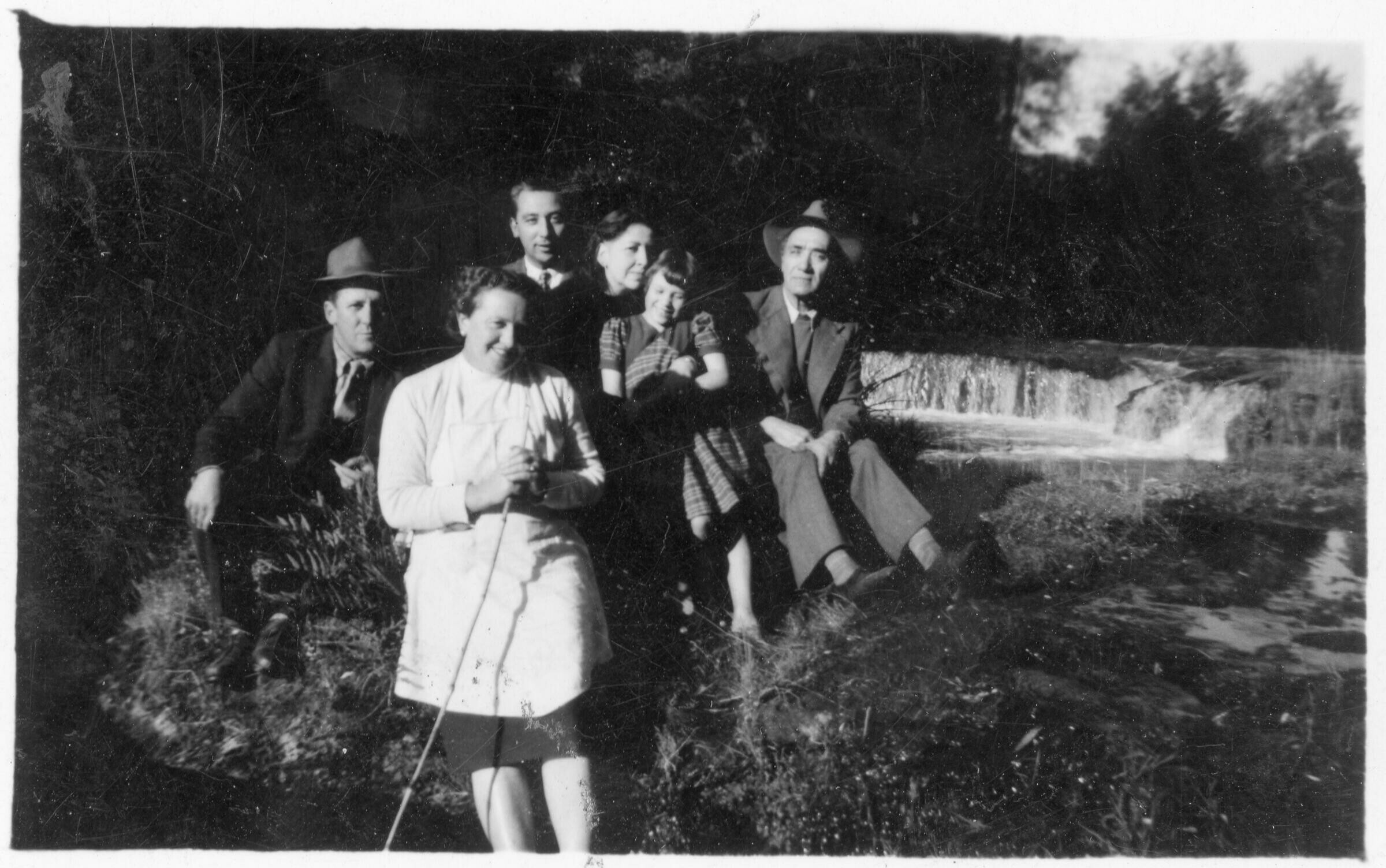 Retrato grupal de plano entero de Hombres, Mujeres y una niña posando sentados en el pasto. En el fondo hay una pequeña cascada no identificada.