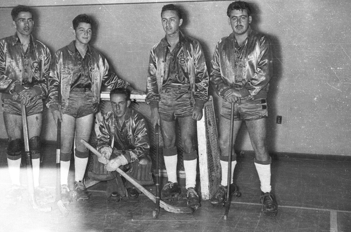 Retrato grupal de plano entero de un equipo de hockey masculino no identificado. En sus poleras tienen grabadas las iniciales CB. están formados junto al arco sobre la cancha en el interior de un gimnasio