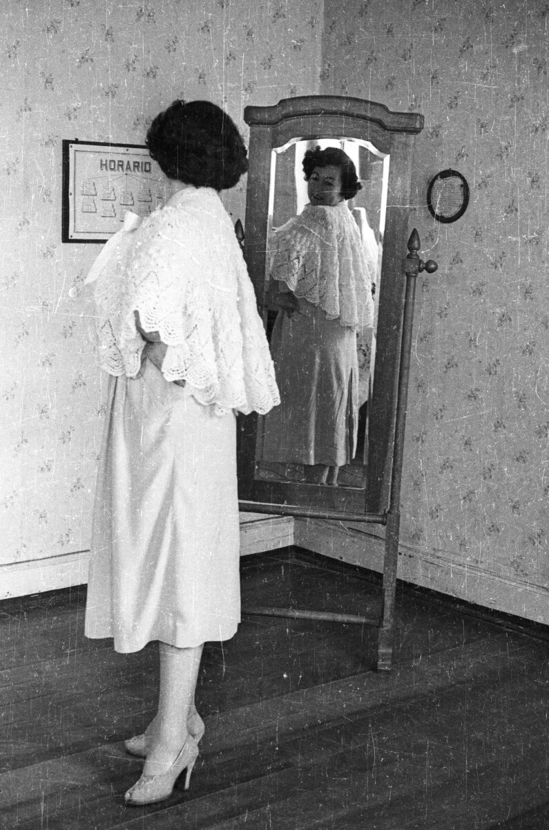 Plano entero de una mujer mirando como le queda un chal tejido a crochet en un espejo de cuerpo entero en el interior de una casa.