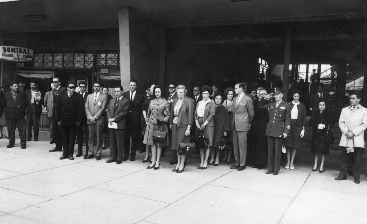 plano general de un grupo de personas paradas sobre la vereda. En el medio se encuentra Eva Sámano, primera dama de México y Olga Boettcher tomadas del brazo. Se encuentran en el frontis de Correos de Chile, durante un homenaje realizado para la primera dama durante su visita a la ciudad de La Unión.