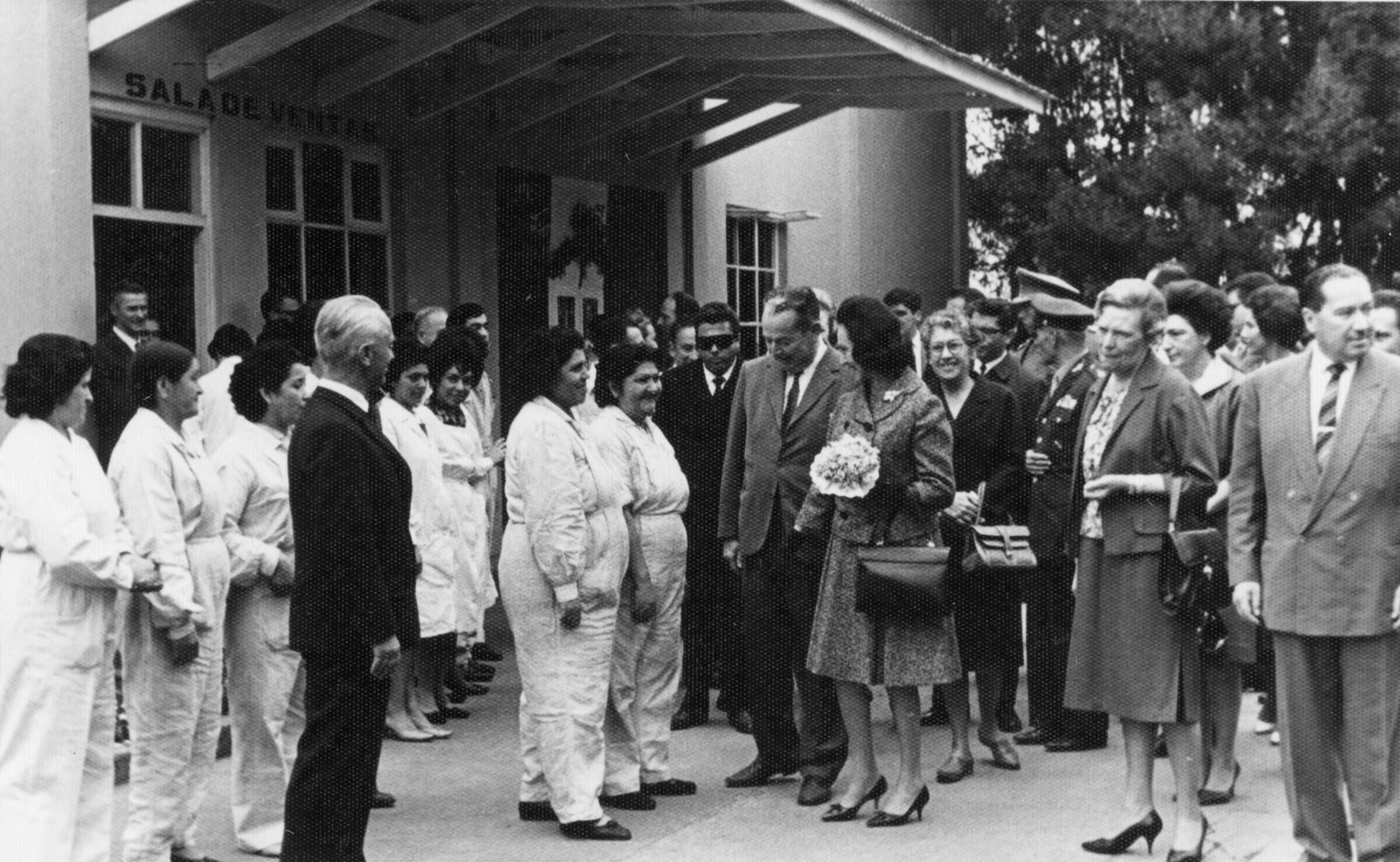 -plano general de Eva Sámano de López Mateos, primera dama de México, conversando con un grupo de trabajadoras a un costado del edificio, el cual tiene un letrero que dice --sala de ventas--. A un costado la acompaña Olga Boettcher, quien tiene una cartera en la mano. En el fondo, un grupo de Hombres y Mujeres las acompañan durante la visita a la planta de Lino de La Unión-