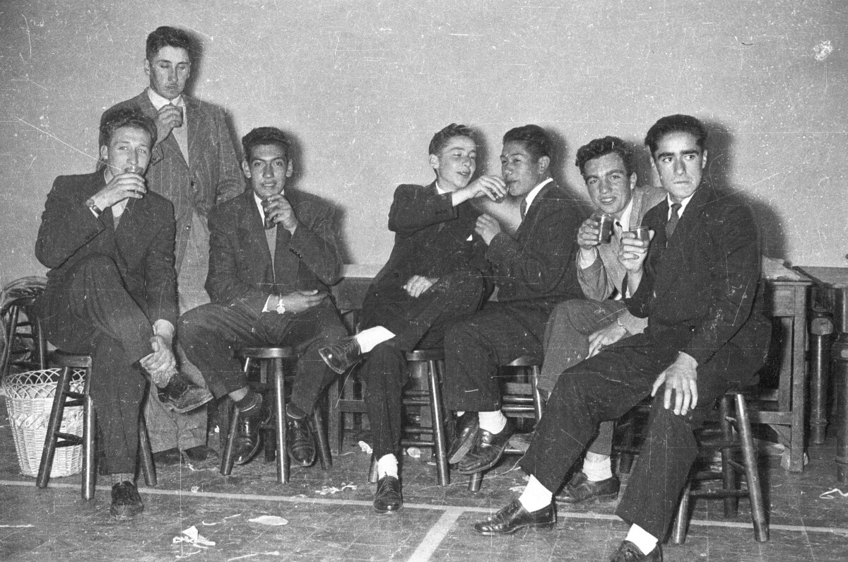 plano general de un grupo de Hombre Joven vestidos de traje posando con una copa en sus manos mientras están sentados sobre unos bancos. Están en el interior de un gimnasio durante una fiesta.