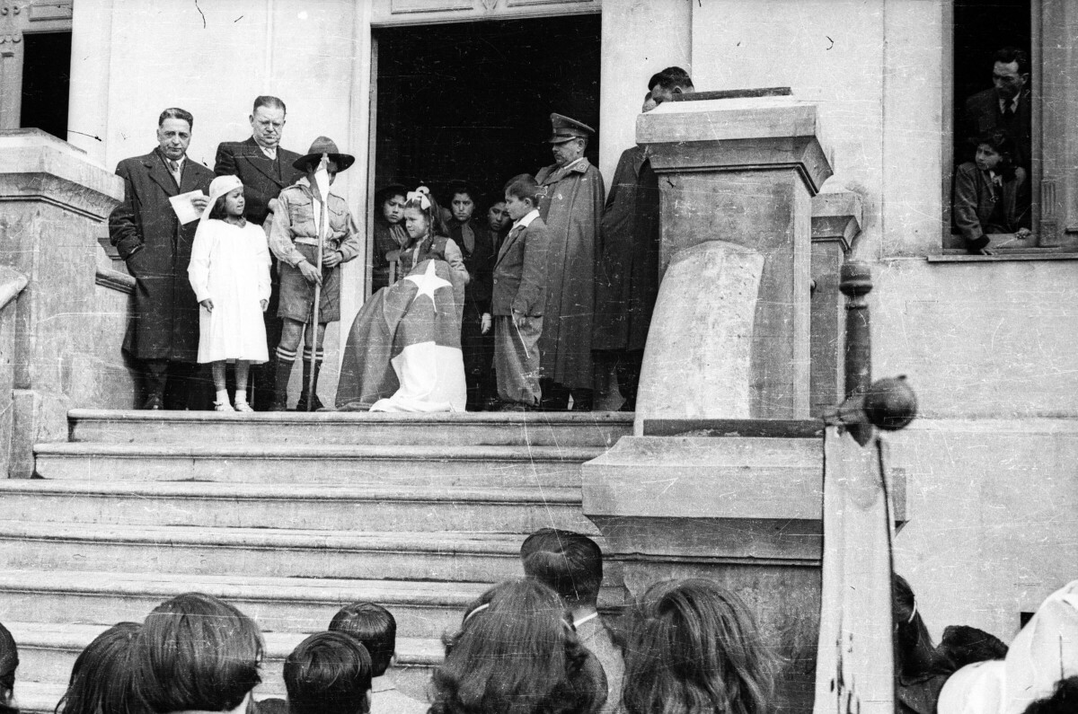 plano general de un niña sosteniendo una bandera chilena junto a otra Niña perteneciente a la Cruz Roja Juvenil de la Escuela Nº2 y un niño perteneciente a una brigada de scout. están junto a un grupo de hombres vestidos de traje y otro usando un uniforme militar en las afueras de la Municipalidad de La Unión.