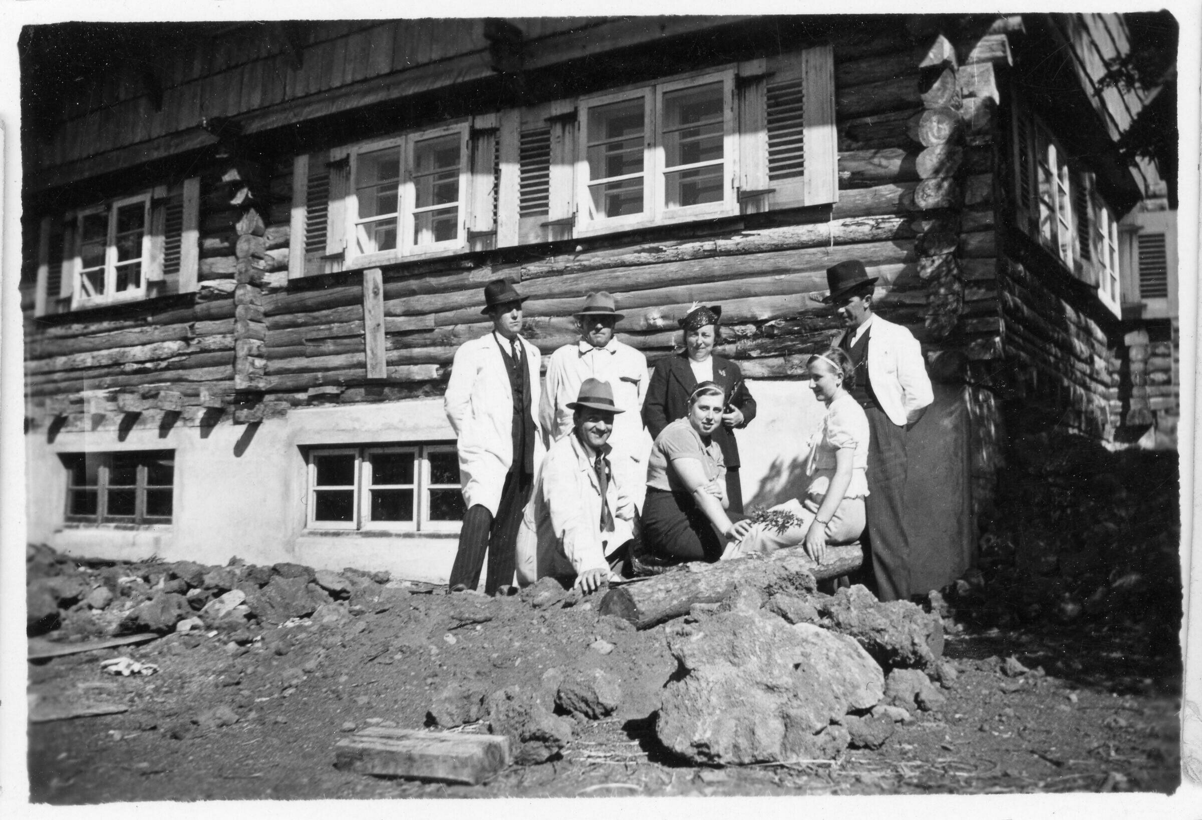 Retrato grupal de plano entero de hombres y mujeres posando en el frontis de una casa de madera, en un lugar no identificado.
