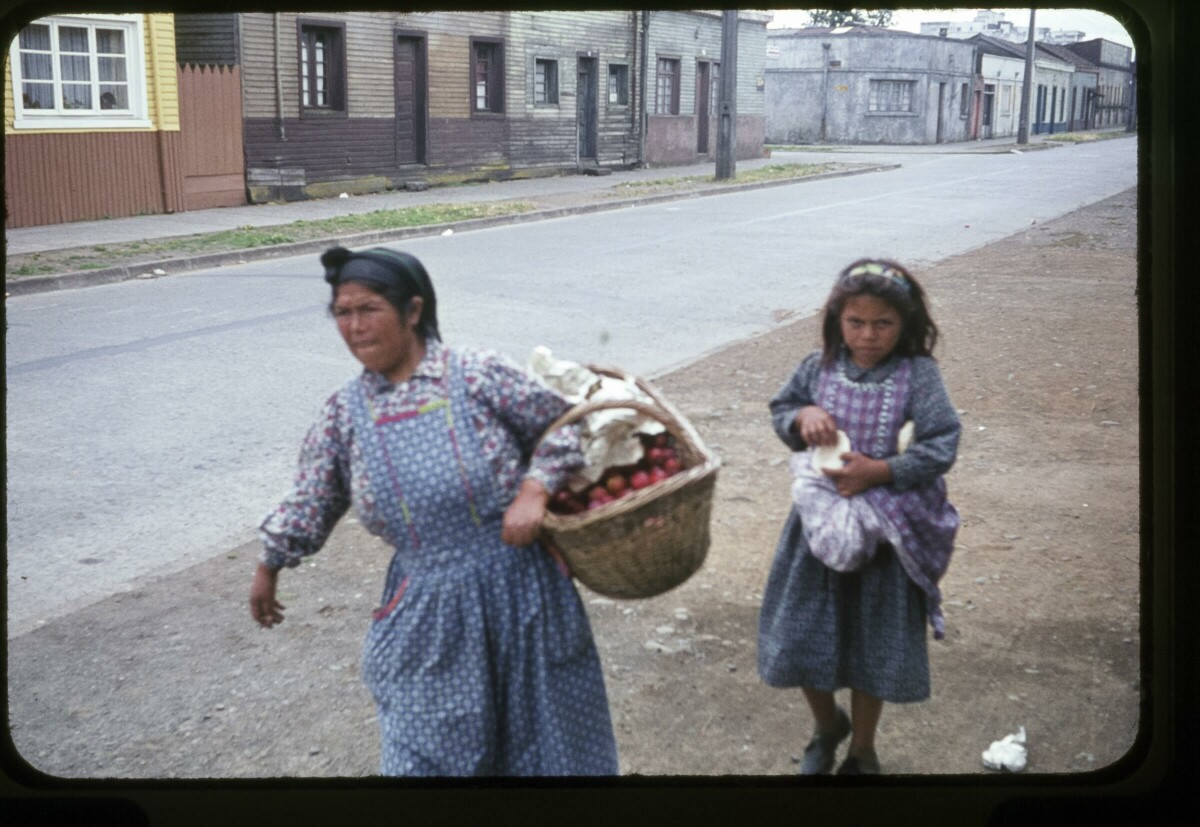 Retrato plano americano de mujer con delantal vendiendo fruta en un canasto de mimbre, a su lado una niña con vestido y delantal, caminando por una calle