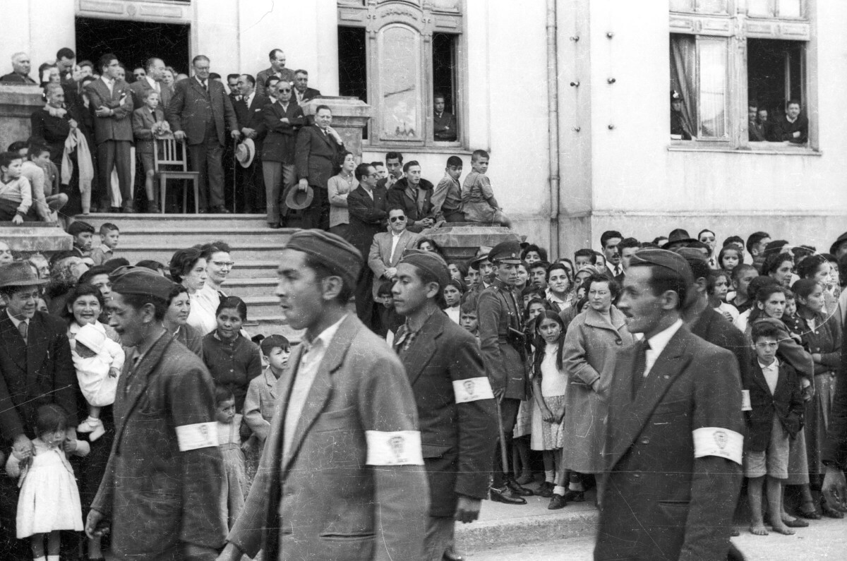 Plano entero de un grupo de Hombres, desfilando con una cinta amarrada en el brazo. En el fondo, se encuentra Carlos Vogel junto a una silla observandolos y también un grupo numeroso de personas en el frontis de la municipalidad