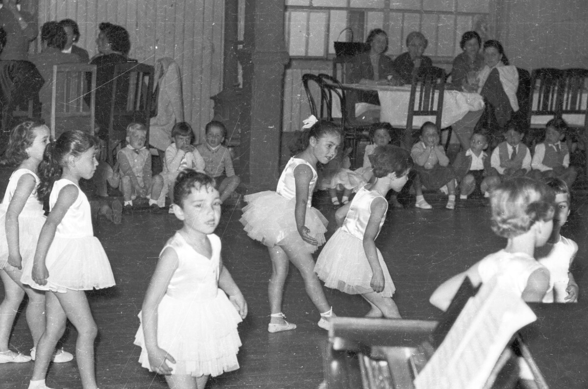 plano general de un grupo de niñas, caracterizadas como bailarinas de ballet, realizando una presentación artística. En el fondo, un grupo de niñas y Niños se encuentran sentados en una hilera observándolas. Se encuentran en una estructura no identificada