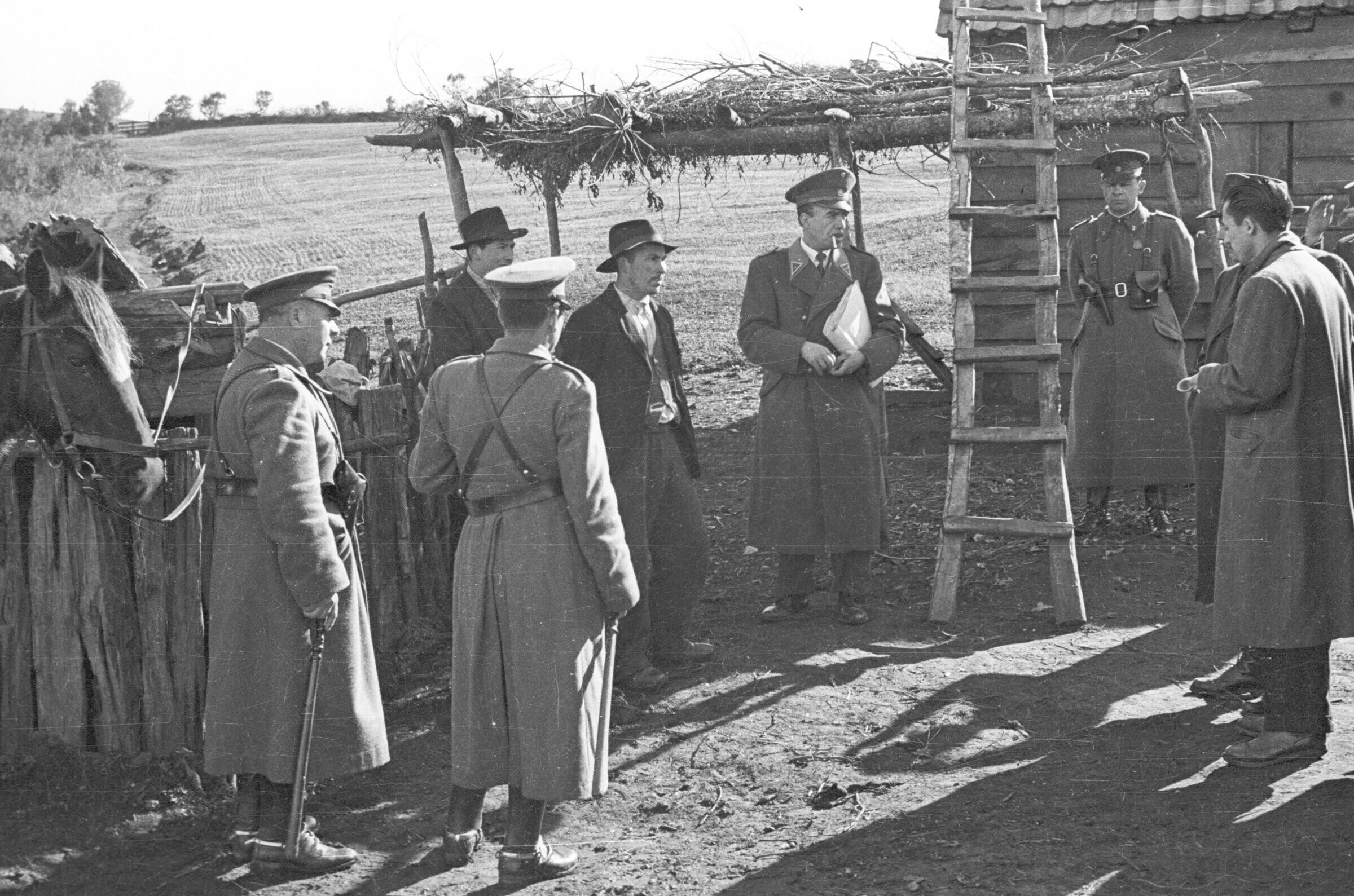 plano general de un grupo de carabineros reunidos en compañía de dos hombres con sombrero y otros dos hombres de abrigo, en el patio de una casa rural, en la cual están recreando un asalto. La casa está ubicada en un territorio no identificado