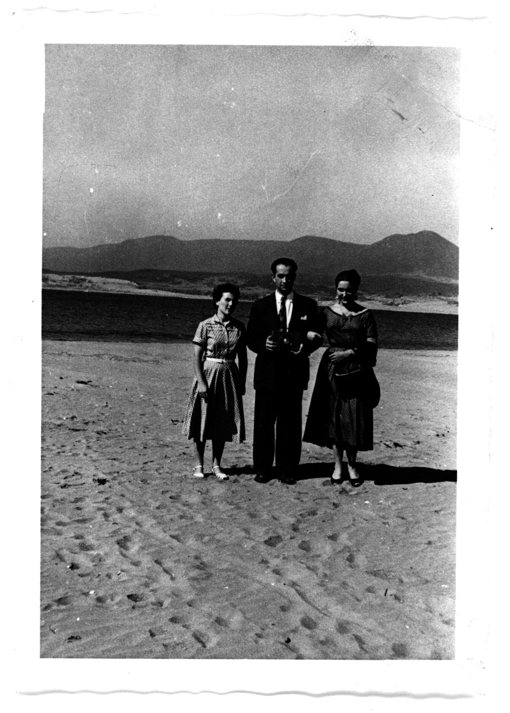 Dos mujeres en vestido y un hombre de terno sosteniendo una cámara, de pie sobre la arena en Pichidangui