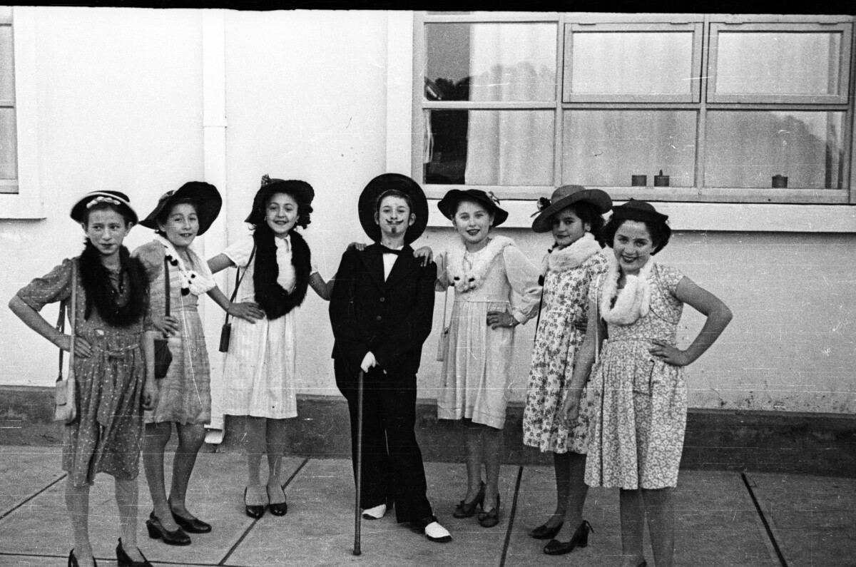 Retrato grupal de Plano entero de niñas disfrazadas de damas antiguas, una de ellas está disfrazada de hombre y lleva dibujado un bigote. Están posando en las afueras de un colegio