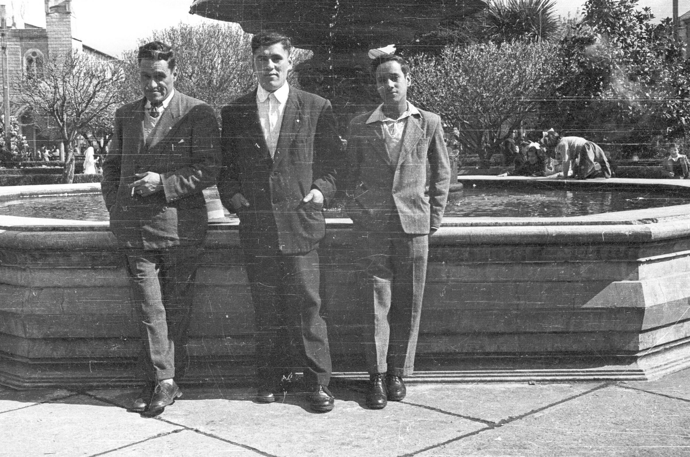 Plano entero de tres hombres posando de pie vestidos de traje, uno de ellos está fumando, tiene un cigarro en la mano y la otra mano en el bolsillo de su pantalón. están posando junto a la pileta en el interior de la Plaza de La Concordia en La Unión