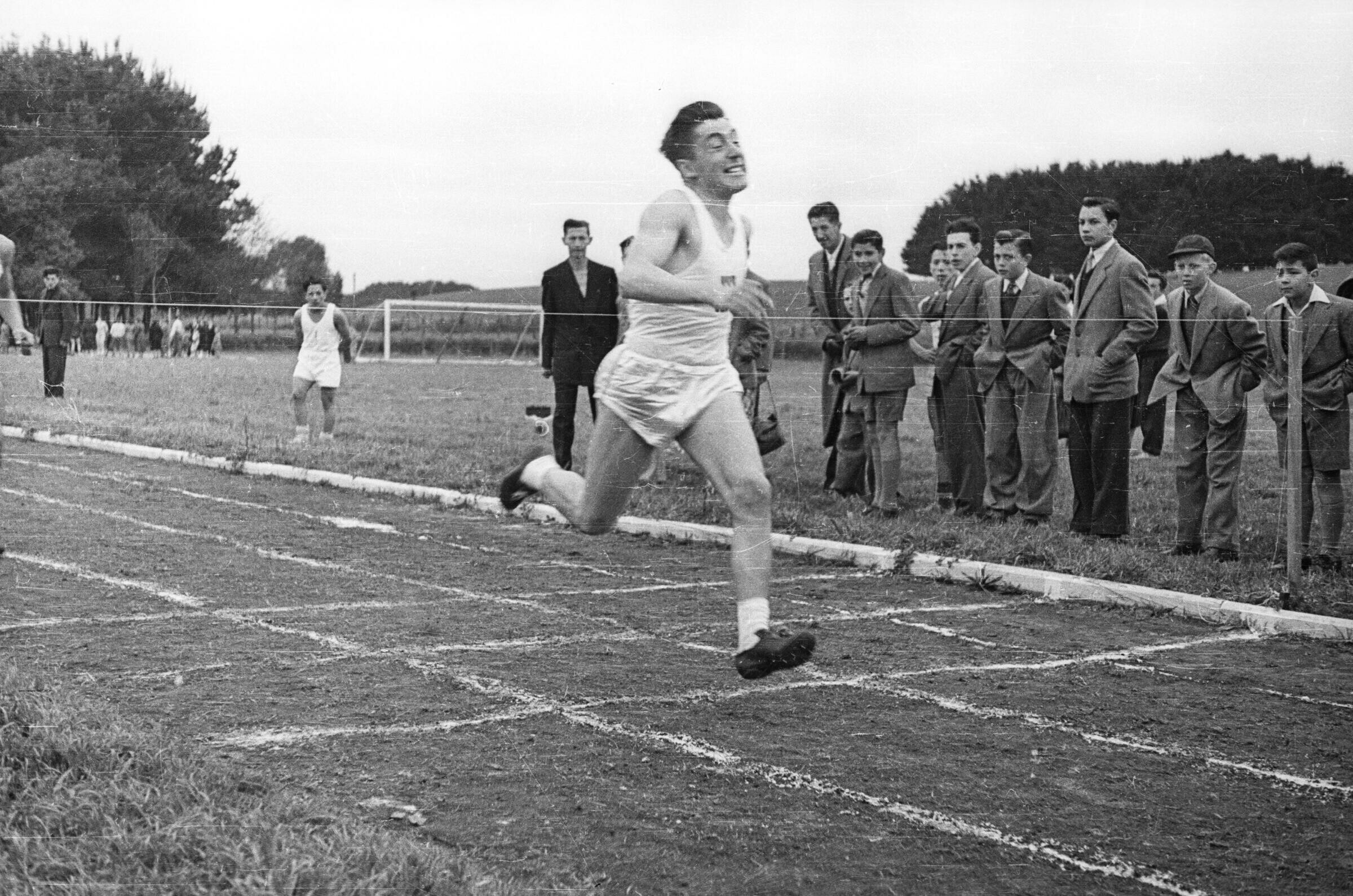 Plano entero de un hombre llegando a la línea de meta durante una carrera, a su alrededor un grupo de Niños y Hombres lo observan de pie junto a la pista durante una jornada de competiciones de juegos olímpicos