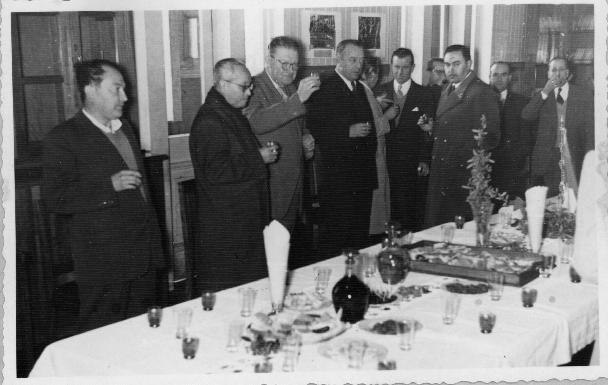 plano general de un grupo de hombres brindando con una copa en la mano. En el medio, se encuentra Olga Boettcher, gobernadora de La Unión, reunida con un hombre y un carabinero. Están ubicados detrás de una mesa, en el interior de una infraestructura no identificada, durante un banquete