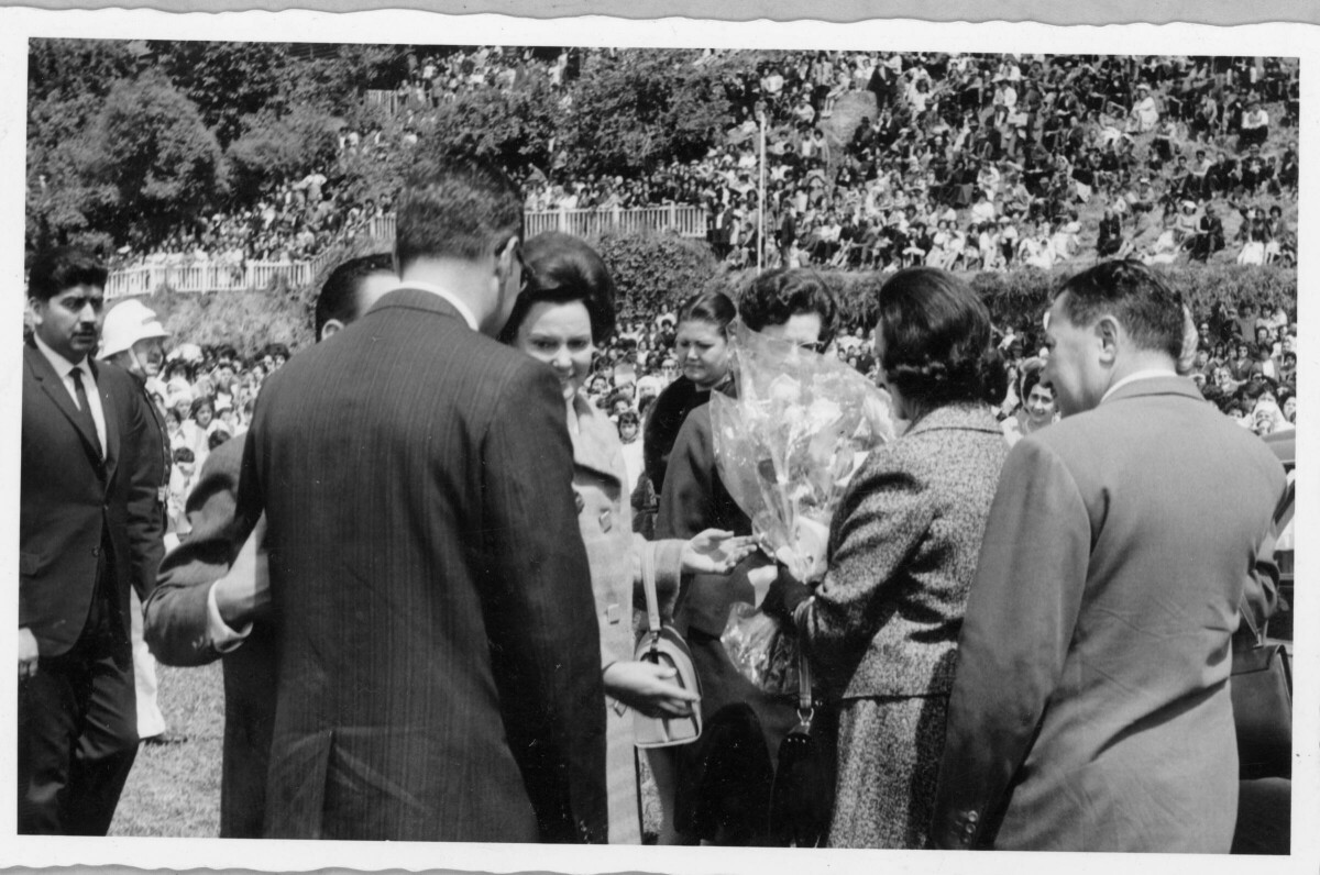 plano general de un grupo de mujeres y hombres reunidos sobre una cancha, conversando. La mujer que está de espaldas a la cámara con un ramo de flores en las manos es Eva sámano, durante su visita a La Unión. Se encuentran en el interior del estadio Carlos Vogel durante un homenaje realizado en su honor