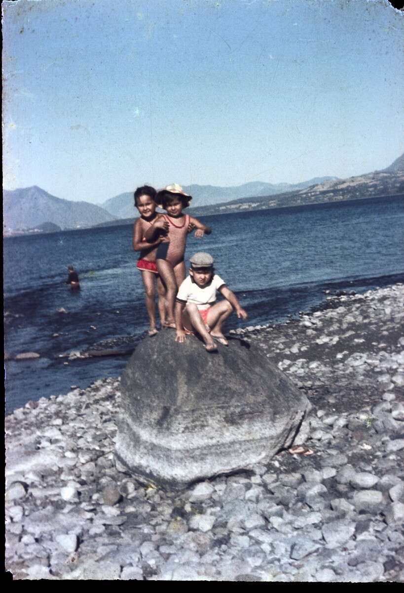 Retrato de plano entero de dos niños y una niña posando encima de una piedra a orilla de un lago no identificado