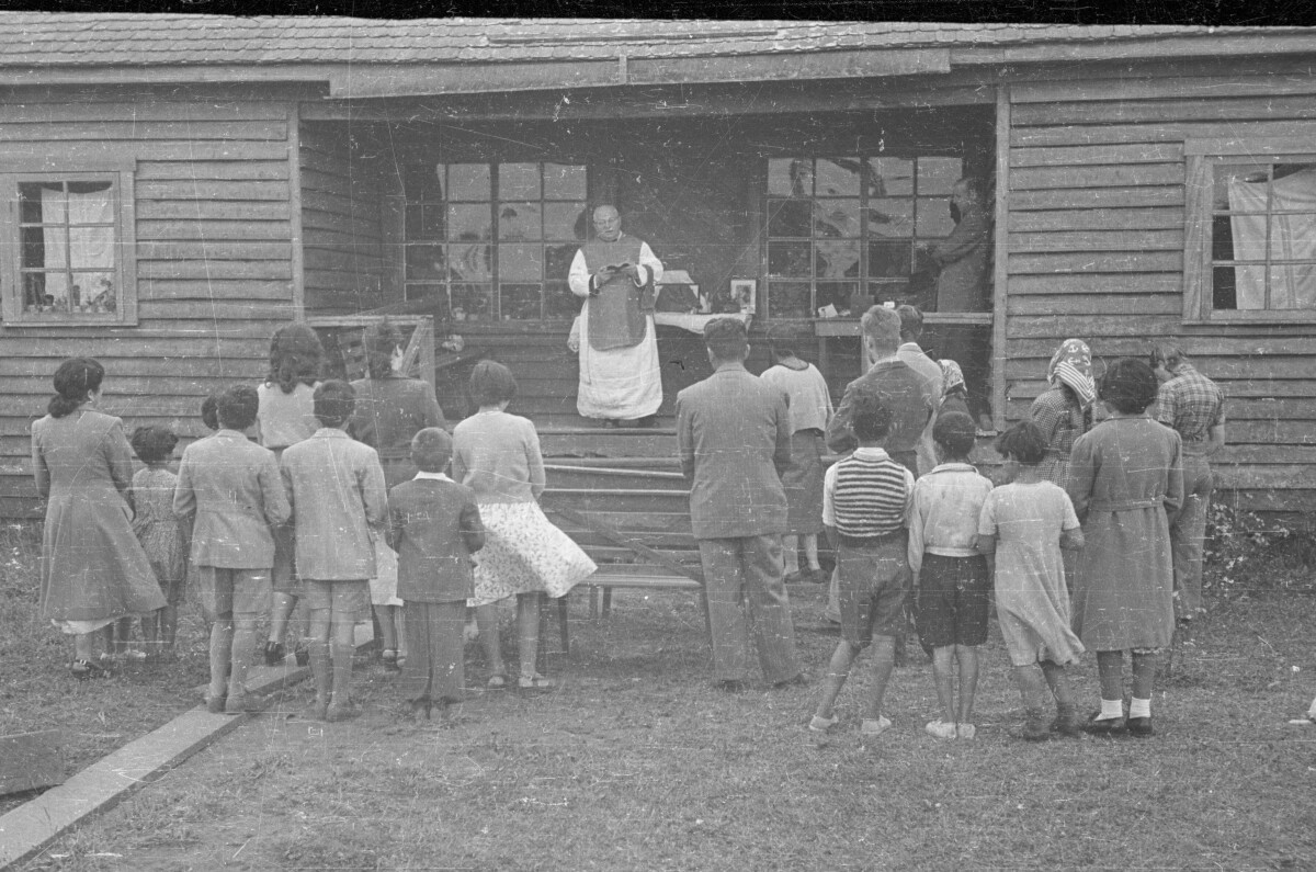 Plano general de hombres, mujeres y niños reunidos de pie frente a sacerdote en las afueras de una casa durante una misa católica.
