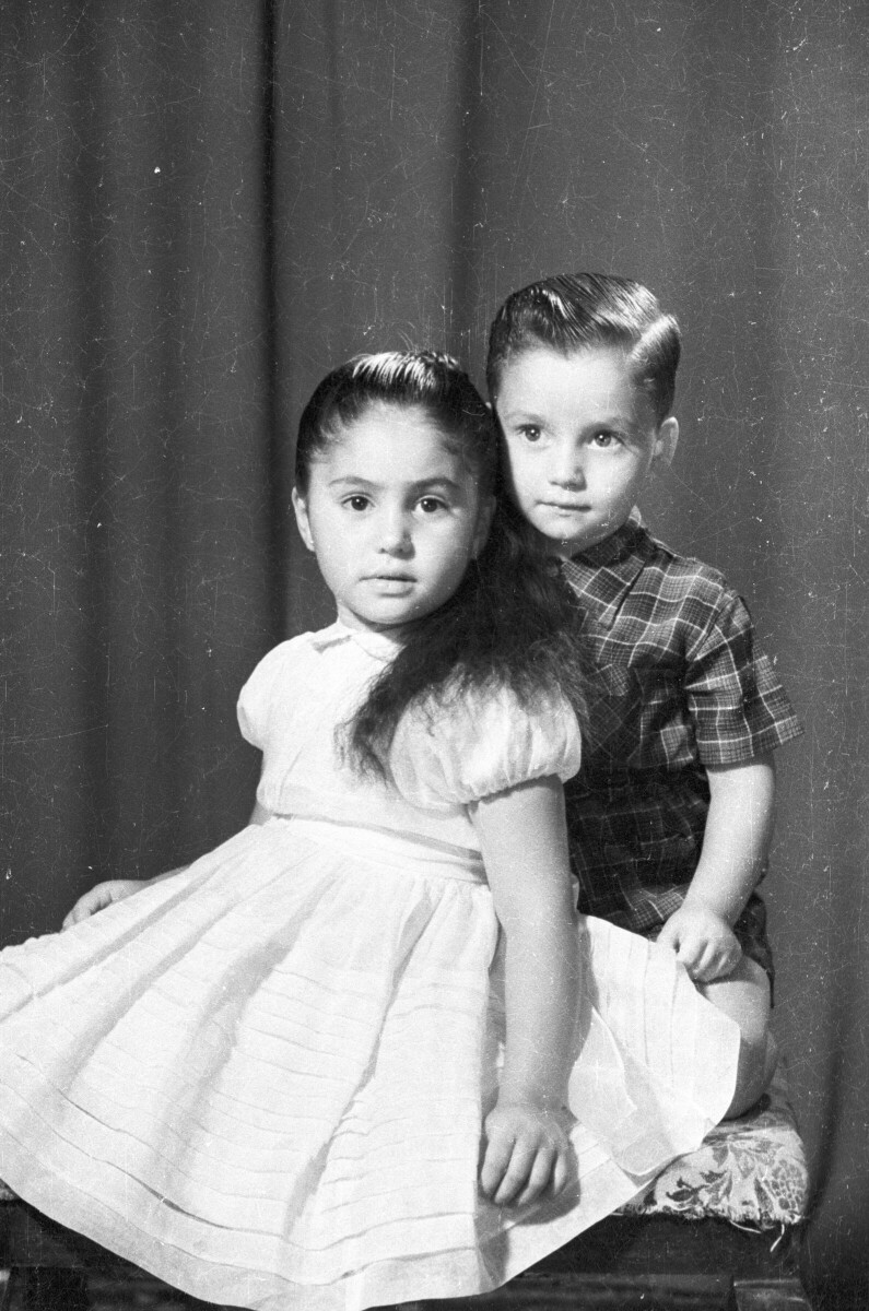 Retrato de estudio Plano americano de niña con Vestido sentada en mesa apoyada sobre no con Traje que está arrodillado en la mesa.