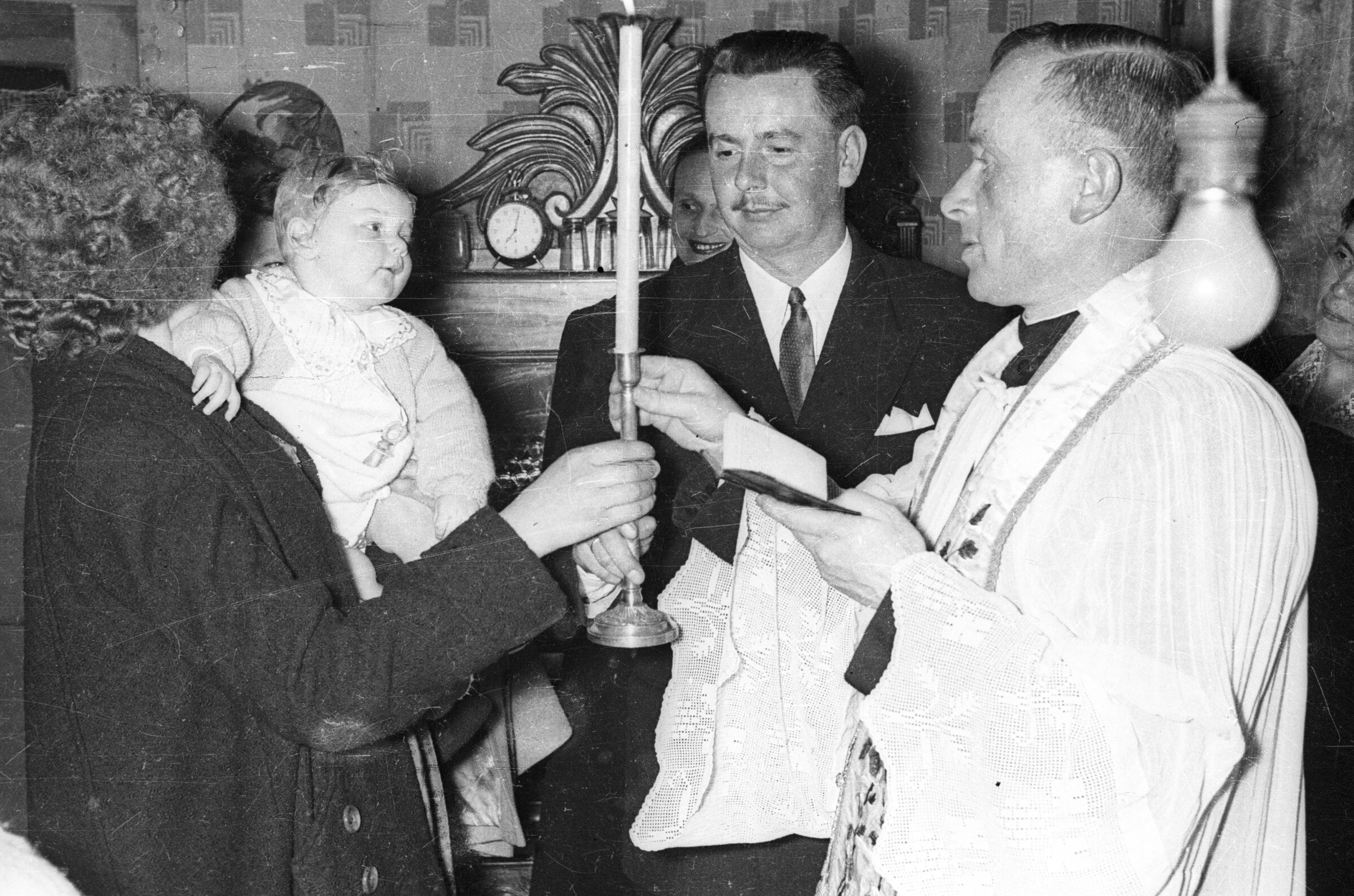 Plano medio largo de un sacerdote entregándole una vela a una mujer quien tiene en brazos a una Bebé. La vela también la está recibiendo el hombre vestido de Traje que los acompaña durante el bautizo de la Niña realizado en el interior de una casa