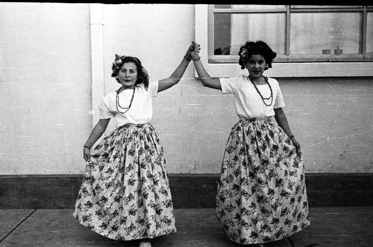 Retrato de plano entero de dos niñas tomadas de la mano vestidas con una falda ancha y una polera blanca, ambas llevan un cintillo con flores y collares, están posando en el patio de un colegio